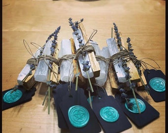 May include: A collection of bundled sage and selenite sticks with lavender sprigs, tied with twine. The bundles are arranged on a wooden surface with black tags featuring a pentagram seal in teal.