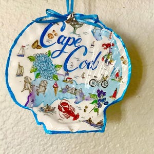 May include: A decorative scallop shell ornament featuring the hand-painted text Cape Cod in blue script. The surface displays colorful illustrations of sailboats, a lobster, a bicycle, lighthouses, hydrangeas, and an ice cream cone, finished with a blue ribbon.