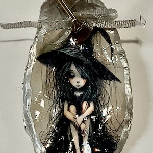 May include: A small, silver-colored, seashell-shaped ornament with a black and white illustration of a witch sitting with her legs crossed. The witch is wearing a black dress and a black hat. A small gold broom is attached to the top of the ornament.