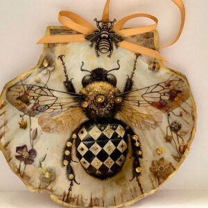 May include: A decorative seashell featuring a detailed illustration of a bee with a black and white checkered abdomen. The bee has ornate wings and is adorned with a small bee charm and an orange ribbon for hanging. The shell has a gold trim.