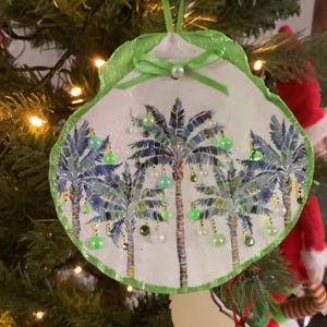 May include: A seashell-shaped Christmas ornament with a green ribbon and pearl accent. The ornament features a palm tree design in blue and green, adorned with green and white beads. It hangs on a Christmas tree with twinkling lights.