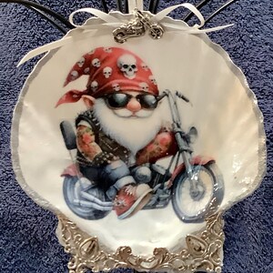 May include: A decorative seashell ornament featuring a biker gnome riding a motorcycle. The gnome wears a red bandana with skull print, sunglasses, and a leather vest. The ornament has a silver trim and a small motorcycle charm.