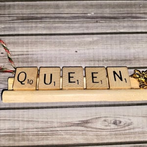 May include: A wooden ornament spelling "QUEEN" with Scrabble tiles, a gold bee charm, and a red and white twine hanger.