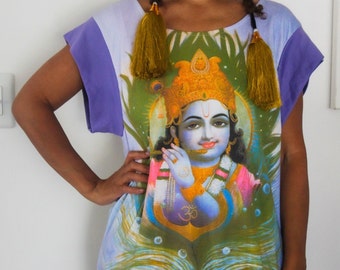 May include: A purple and white t-shirt with a colorful image of a Hindu deity, Krishna, playing a flute. The deity is wearing a yellow crown and a blue and gold necklace. The t-shirt has a purple trim around the neck and sleeves.