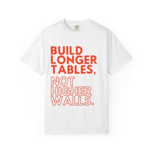 May include: White t-shirt featuring the red text "BUILD LONGER TABLES, NOT HIGHER WALLS." The shirt has a crew neck and short sleeves. The text is in a bold, sans-serif font.