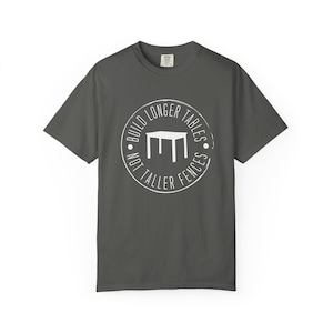 May include: Dark grey t-shirt with a white circular graphic. The graphic features a table illustration and the text "BUILD LONGER TABLES NOT TALLER FENCES". The shirt has short sleeves and a crew neck.