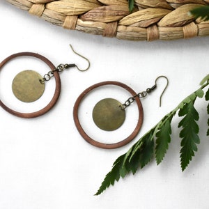 May include: A pair of brown wooden hoop earrings with a gold metal circle pendant hanging from each hoop.