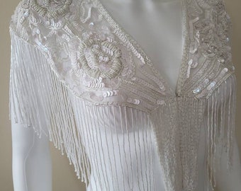 May include: A white beaded shawl with fringe. The shawl has a V-neck and is decorated with sequins and beads.