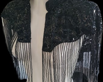 May include: A black sequined bolero jacket with fringe detailing. The jacket has a high collar and is adorned with sequins and long fringe. The garment is displayed on a white mannequin against a black backdrop, showcasing its intricate design and texture.