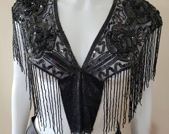 May include: A black beaded and sequined top with a V-neck design. The top features floral embellishments on the shoulders and a fringe detail along the bottom. The garment is displayed on a white mannequin.
