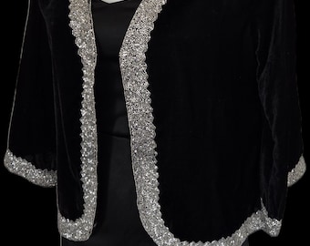 May include: Black velvet jacket with silver sequin trim. The jacket has three-quarter sleeves and a V-neckline. The trim is along the edges of the jacket and sleeves. The jacket is worn over a black dress.