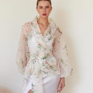 Può includere: Un abito lungo in raso bianco con spacco alto è abbinato a un top avvolgente trasparente con stampa floreale. Il top avvolgente presenta uno scollo a V e maniche lunghe, con una fascia abbinata legata in vita. Una collana d'oro completa il look.