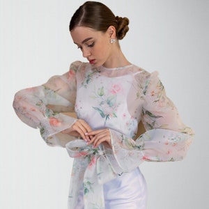 Peut inclure: Une robe blanche à imprimé floral avec des manches bouffantes transparentes. Les manches sont nouées au poignet avec un nœud.