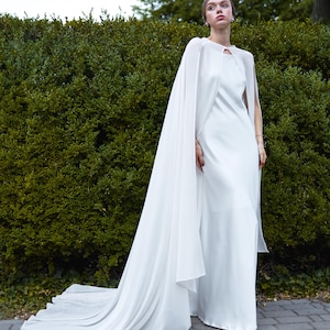 May include: A white wedding dress with a flowing cape. The dress has a keyhole neckline and a long train. The cape drapes over the shoulders and extends to the ground. The dress is made of a soft, flowing fabric.