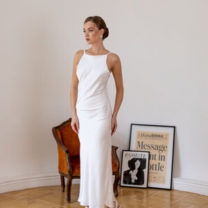 May include: A woman wearing a white, sleeveless, floor-length dress with a scoop neck. She is standing on a wooden floor with a chair and a framed poster behind her. The poster has the text "Le Journal Message nt in bottle" and "Egoiste" on it.