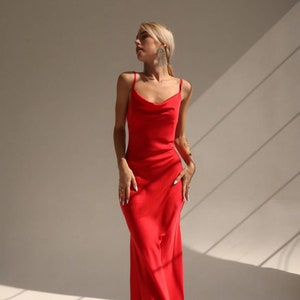 Puede incluir: Una mujer con un vestido maxi de sat&eacute;n rojo con cuello de pico y tirantes finos. Est&aacute; de pie sobre un fondo blanco con luz que entra por una ventana.