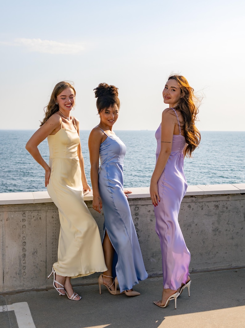 May include: Three women wearing long, silky dresses in yellow, blue, and purple stand on a concrete platform overlooking the ocean. They are all smiling and looking at the camera.