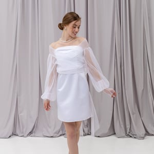 Peut inclure: Une robe blanche &agrave; &eacute;paules d&eacute;nud&eacute;es avec une superposition de tulle et des manches longues. La robe a un corsage ajust&eacute; et une jupe courte. Le mannequin porte la robe avec un collier de perles et des talons nus.