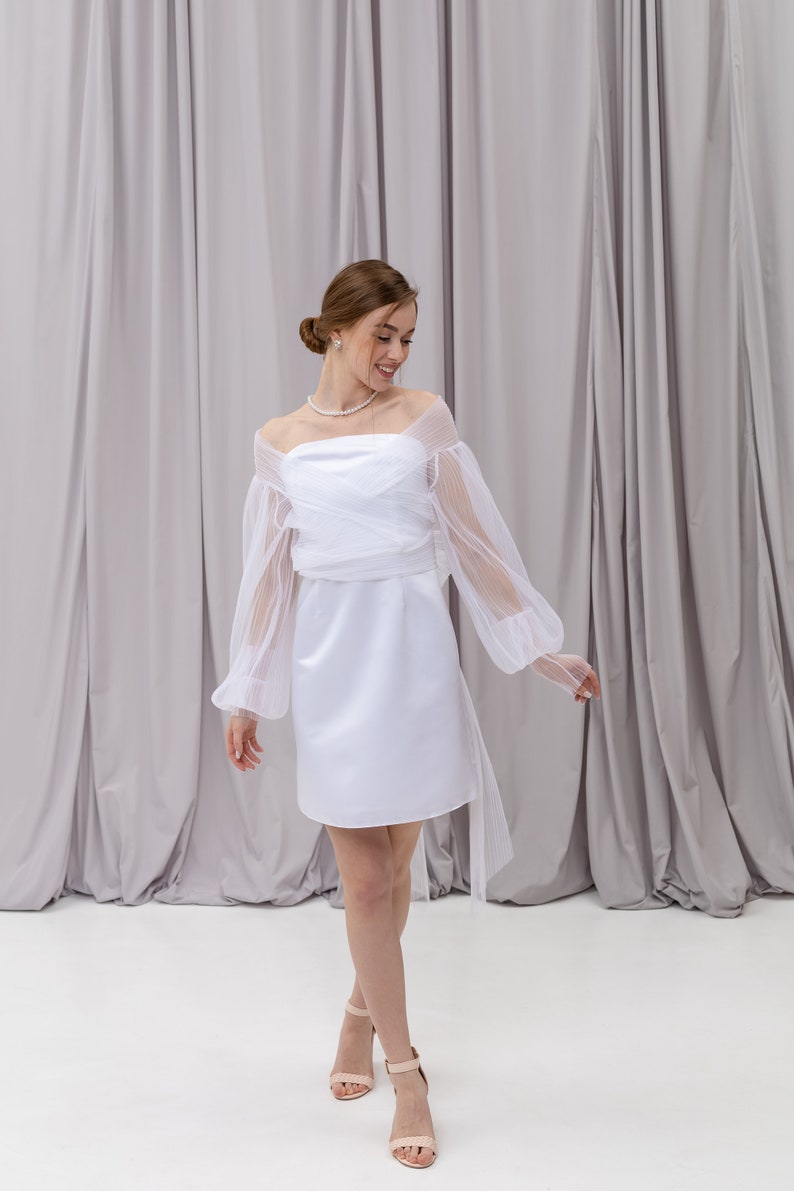 Peut inclure: Une robe blanche &agrave; &eacute;paules d&eacute;nud&eacute;es avec une superposition de tulle et des manches longues. La robe a un corsage ajust&eacute; et une jupe courte. Le mannequin porte la robe avec un collier de perles et des talons nus.