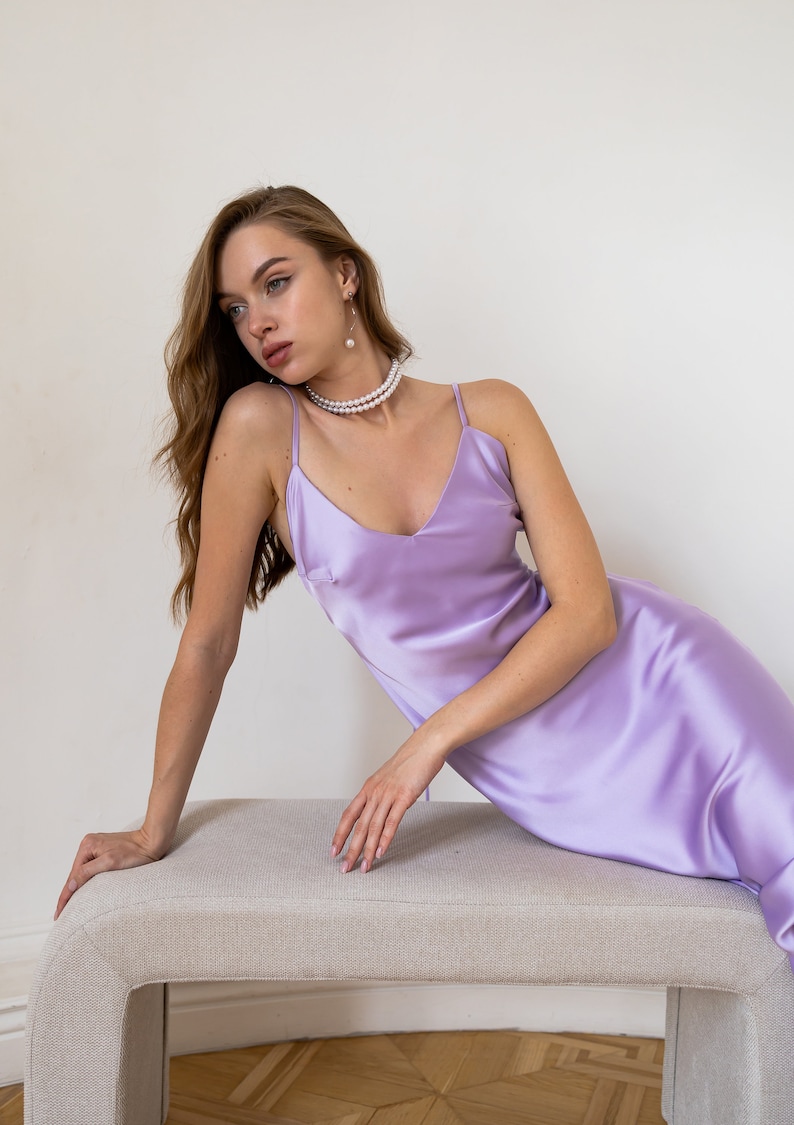 K&ouml;nnte beinhalten: Eine Frau in einem helllila Satin-Schlafkleid mit einer Perlenkette und Ohrringen. Sie sitzt auf einer hellbeigen Bank.