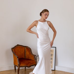 May include: A woman wearing a white satin slip dress with a halter neckline and a fitted silhouette. She is standing in a room with a wooden floor and a brown armchair. She is wearing white heels.