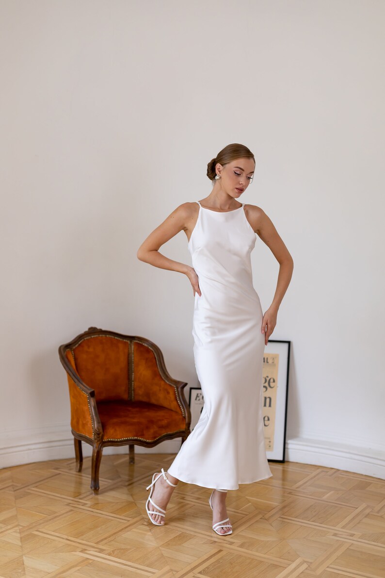 May include: A woman wearing a white satin slip dress with a halter neckline and a fitted silhouette. She is standing in a room with a wooden floor and a brown armchair. She is wearing white heels.