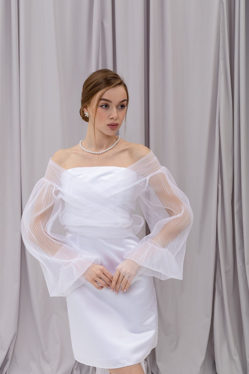 Peut inclure: Une robe blanche &agrave; &eacute;paules d&eacute;nud&eacute;es avec des manches transparentes et bouffantes. La robe a un corsage ajust&eacute; et une jupe fluide. Le mannequin porte un collier et des boucles d'oreilles en perles.