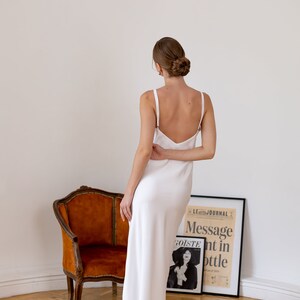 May include: A woman wearing a white, sleeveless, floor-length dress with a deep V-neckline. She is standing in front of a chair and a framed poster with the text "Message in a bottle".