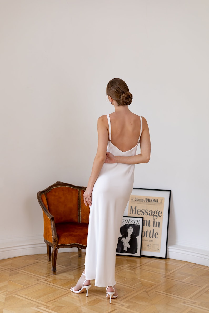 May include: A woman wearing a white, sleeveless, floor-length dress with a deep V-neckline. She is standing in front of a chair and a framed poster with the text "Message in a bottle".