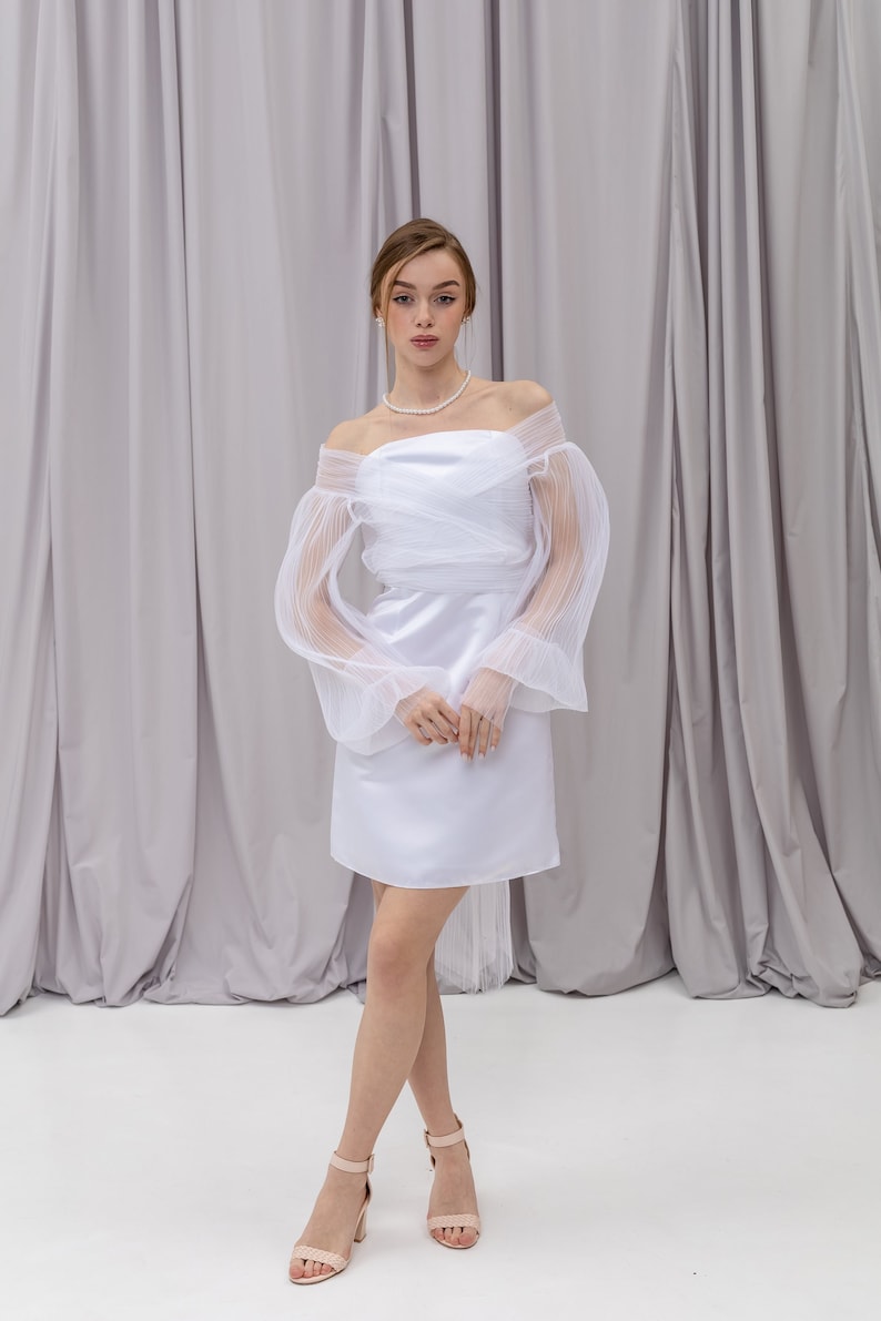 Peut inclure: Une robe blanche &agrave; &eacute;paules d&eacute;nud&eacute;es avec des manches transparentes pliss&eacute;es et une jupe en satin. La robe a un corsage ajust&eacute; et une jupe fluide. Le mannequin porte la robe avec des talons nus.