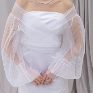 Peut inclure: Une robe blanche &agrave; &eacute;paules d&eacute;nud&eacute;es avec des manches transparentes pliss&eacute;es et un collier de perles.