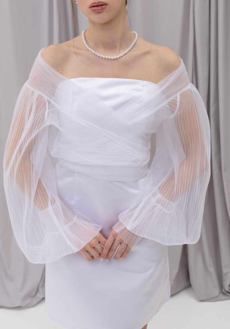 Peut inclure: Une robe blanche &agrave; &eacute;paules d&eacute;nud&eacute;es avec des manches transparentes pliss&eacute;es et un collier de perles.