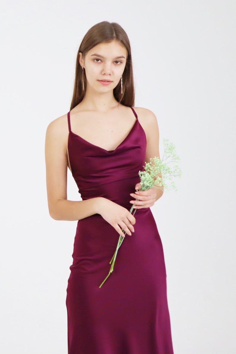 May include: A woman wearing a long, burgundy satin dress with a cowl neck and spaghetti straps. She is holding a bouquet of small white flowers.