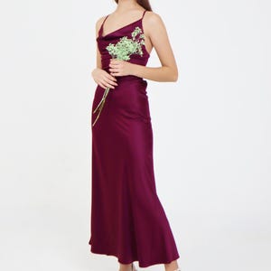May include: A woman wearing a long, burgundy satin dress with a cowl neck and spaghetti straps. She is holding a small bouquet of white flowers and is standing on a white background.