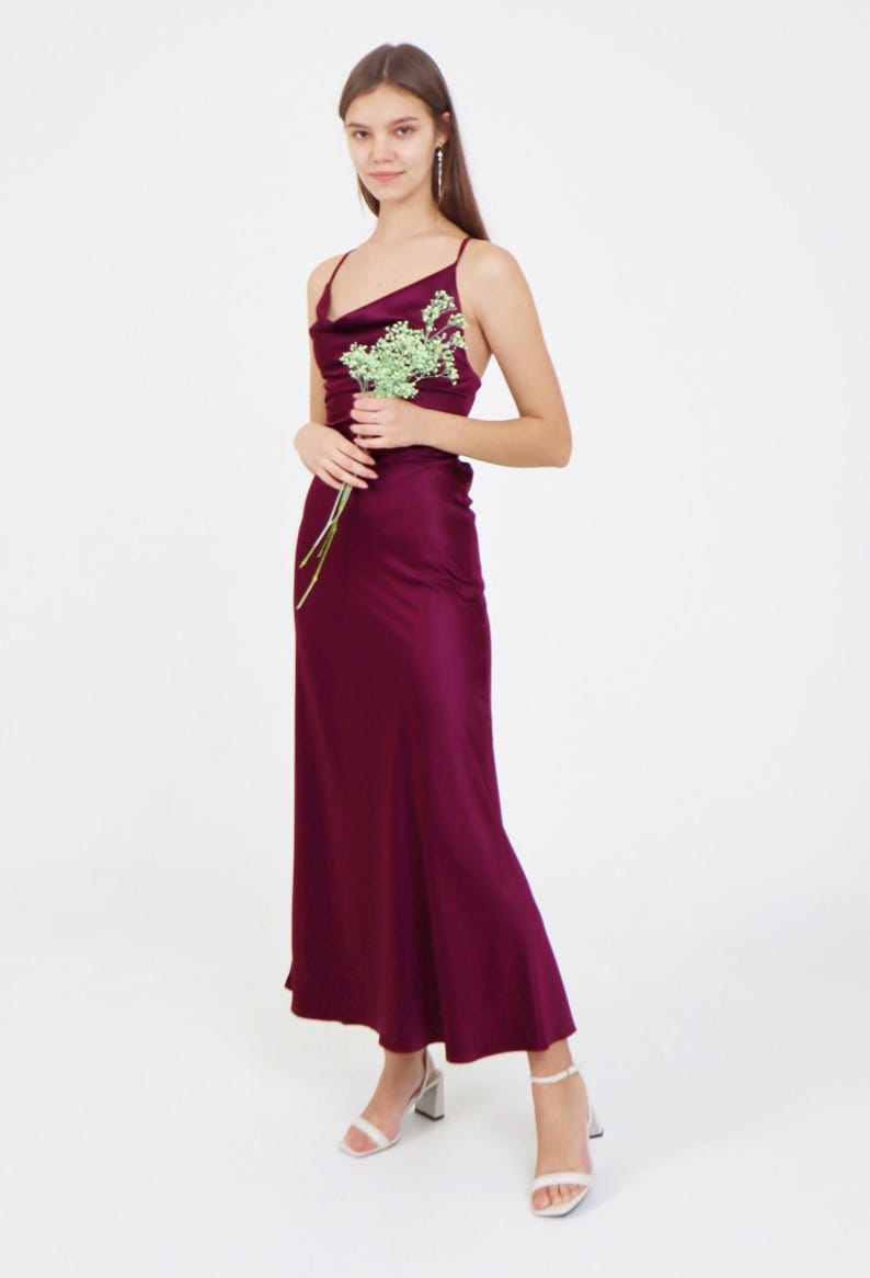 May include: A woman wearing a long, burgundy satin dress with a cowl neck and spaghetti straps. She is holding a small bouquet of white flowers and is standing on a white background.