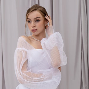 Peut inclure: Une femme portant une robe blanche avec des manches transparentes et bouffantes. Elle se tient devant un fond blanc et a la main dans les cheveux.