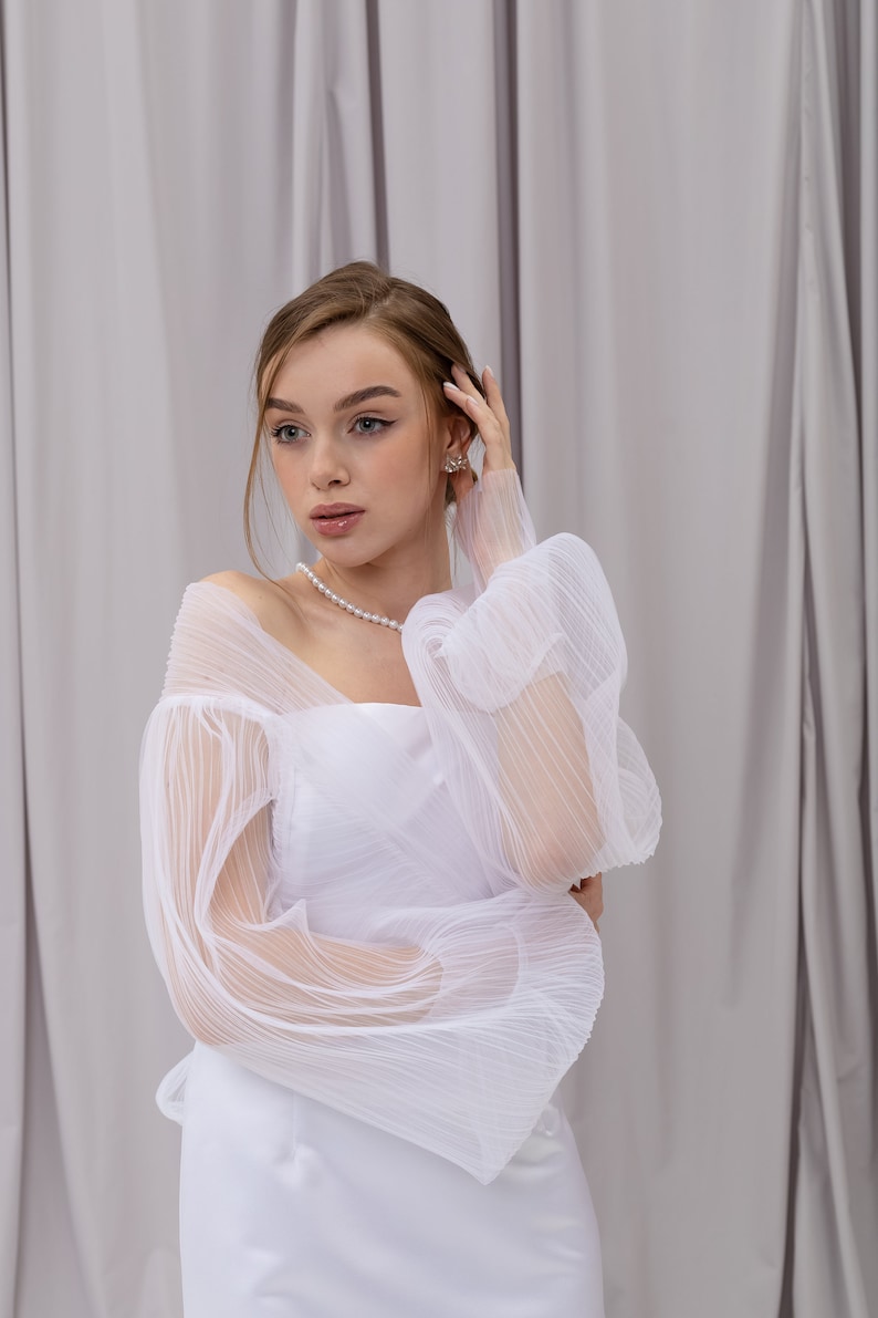 Peut inclure: Une femme portant une robe blanche avec des manches transparentes et bouffantes. Elle se tient devant un fond blanc et a la main dans les cheveux.