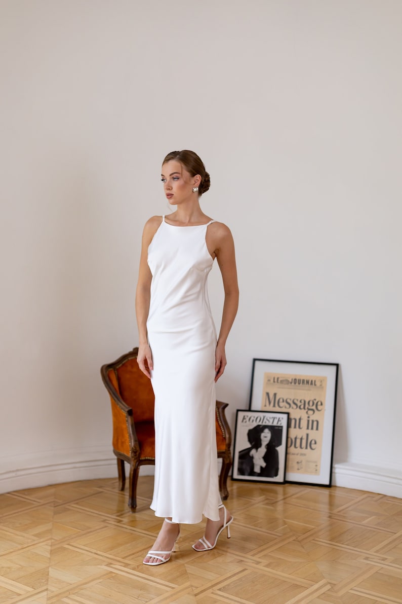 May include: A woman wearing a white, sleeveless, floor-length dress with a scoop neck. She is standing on a wooden floor with a chair and a framed poster behind her. The poster has the text "Le Journal Message nt in bottle" and "Egoiste" on it.