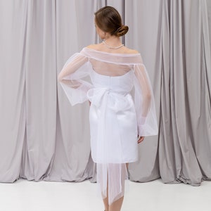 Peut inclure: Une robe blanche &agrave; &eacute;paules d&eacute;nud&eacute;es avec une superposition transparente et un n&oelig;ud dans le dos. La robe a un corsage ajust&eacute; et une jupe fluide.