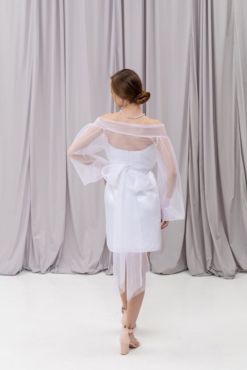 Peut inclure: Une robe blanche &agrave; &eacute;paules d&eacute;nud&eacute;es avec une superposition transparente et un n&oelig;ud dans le dos. La robe a un corsage ajust&eacute; et une jupe fluide.