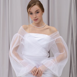 Peut inclure: Une robe blanche &agrave; &eacute;paules d&eacute;nud&eacute;es avec une superposition de tulle &agrave; volants et des manches longues. La robe a un design simple et &eacute;l&eacute;gant et est parfaite pour un mariage ou une autre occasion sp&eacute;ciale.