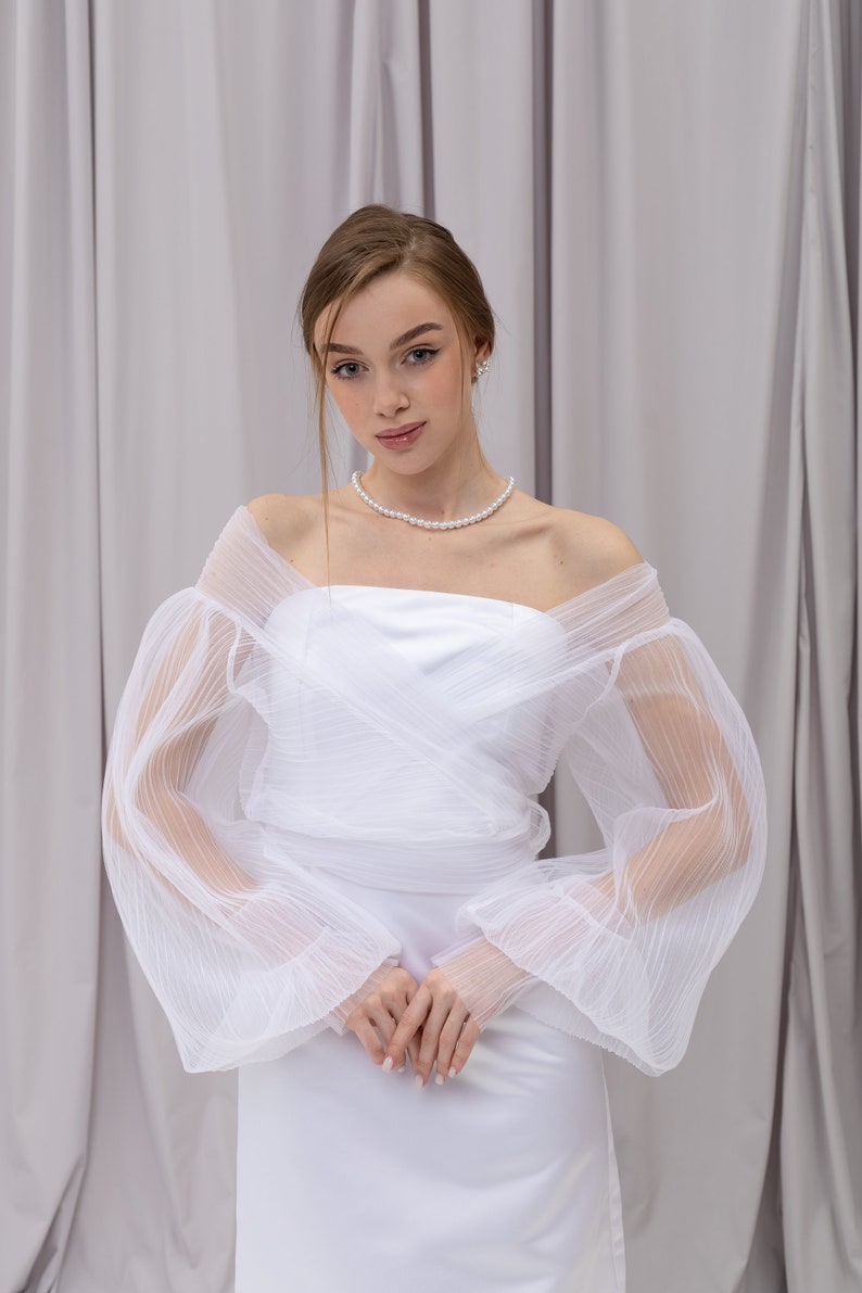Peut inclure: Une robe blanche &agrave; &eacute;paules d&eacute;nud&eacute;es avec une superposition de tulle &agrave; volants et des manches longues. La robe a un design simple et &eacute;l&eacute;gant et est parfaite pour un mariage ou une autre occasion sp&eacute;ciale.