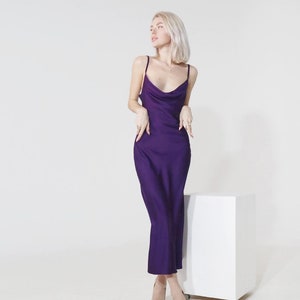 May include: A woman wearing a purple satin slip dress and nude heels stands in front of a white cube.