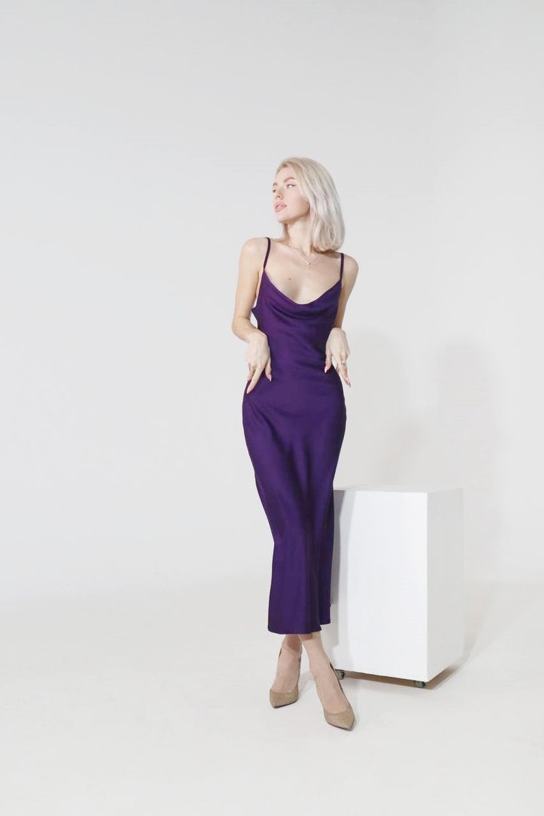 May include: A woman wearing a purple satin slip dress and nude heels stands in front of a white cube.