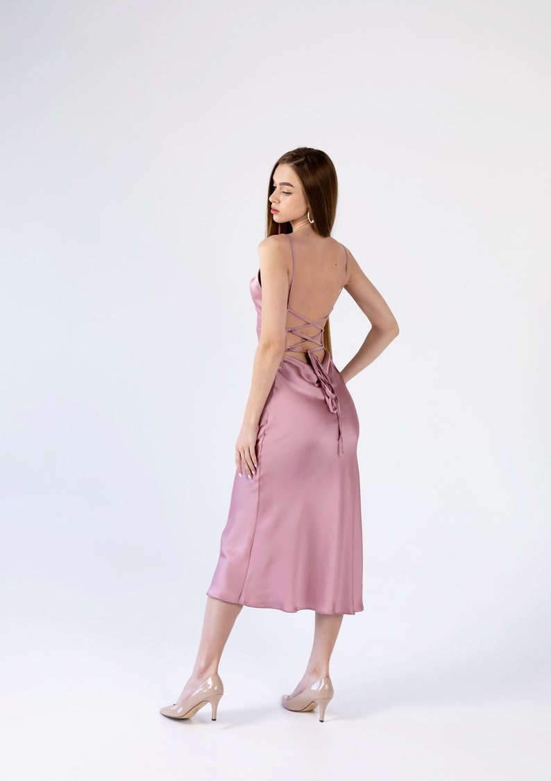 May include: A woman wearing a dusty rose satin midi dress with a lace-up back detail. She is standing on a white background and wearing nude heels.
