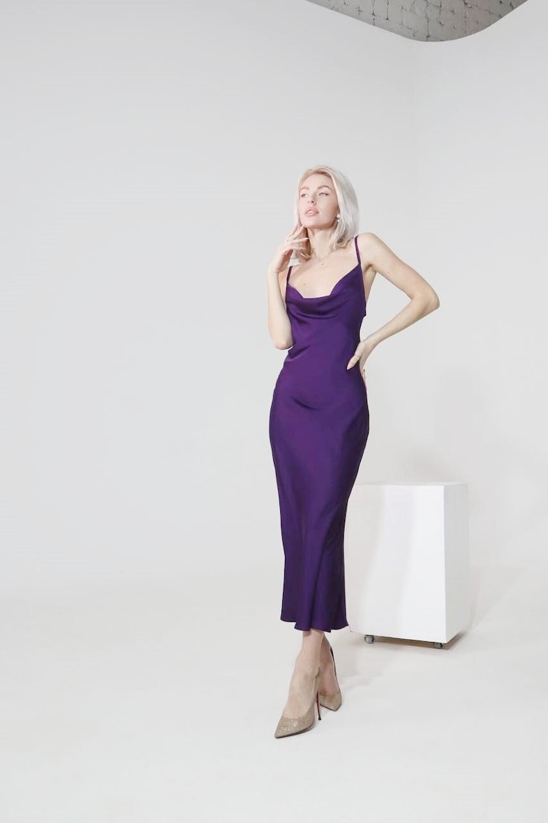 May include: A woman wearing a purple satin cowl neck midi dress and gold sparkly heels. She is standing in front of a white background.