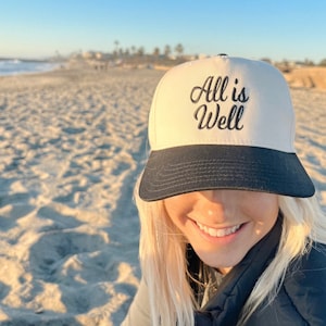 May include: A person wearing a white and black baseball cap with the text "All is Well" embroidered on the front. The person is smiling and has blonde hair. The background is a sandy beach.