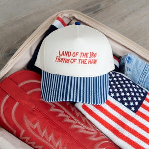 May include: A suitcase containing a white baseball cap with a blue and white striped brim and the text "LAND OF THE YEE HOME OF THE HAW" in red. Also visible are red cowboy boots and a red, white, and blue striped American flag.