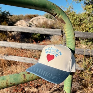 以下が含まれることがあります： 青と白のベースボールキャップに、青い文字で「HEAVEN MADE」と、中央に赤いハートが描かれています。キャップは緑色の金属製のフェンスの上に置かれています。