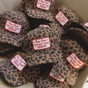 Könnte beinhalten: Eine Ansammlung von Cord-Baseballkappen mit Leopardenmuster. Jede Kappe hat einen rechteckigen Aufnäher mit dem Text "News Flash IT'S NOT THAT SERIOUS" in Rot und Weiß.
